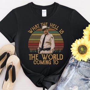 May include: Black t-shirt featuring a vintage-style graphic of a man in a sheriff's uniform against a sunset backdrop. The text reads "WHAT THE HELL IS THE WORLD COMING TO". The shirt is paired with denim shorts and sandals.