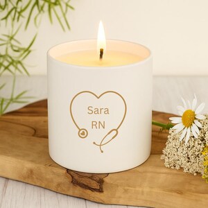 May include: A lit white candle with a gold heart-shaped design, stethoscope, and the text "Sara RN". The candle sits on a wooden board with flowers. The candle's flame is bright against the white ceramic.