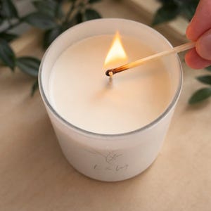 以下が含まれることがあります： 円筒形のガラス容器に入った白いキャンドルで、芯に火が灯っています。キャンドルのワックスは滑らかで無地の白です。木製のマッチ棒が芯に当てられ、明るいオレンジ色の炎が出ています。ガラスには小さく薄い植物のロゴが描かれています。