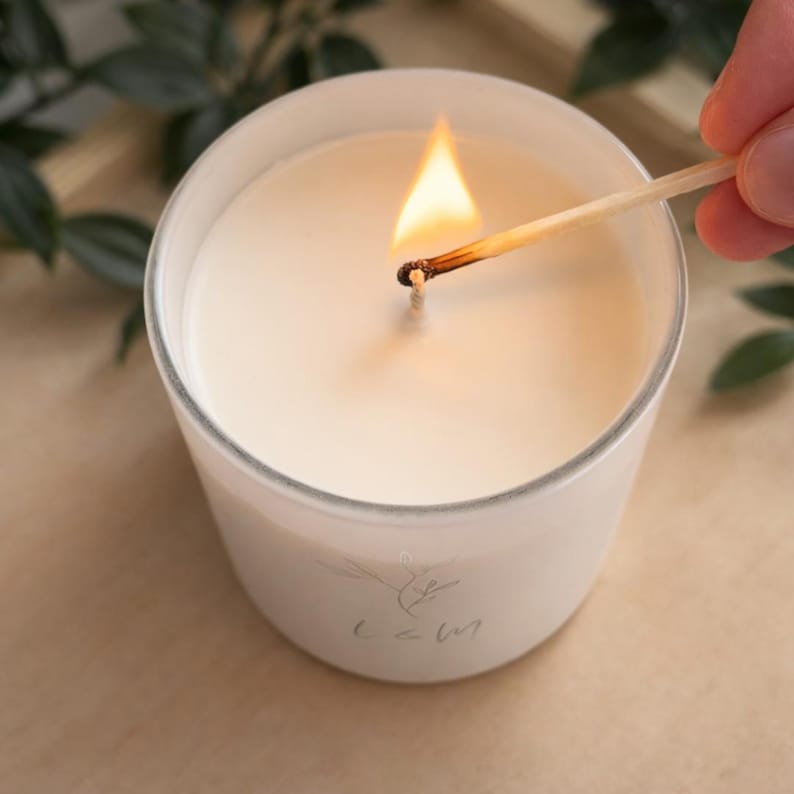 以下が含まれることがあります： 円筒形のガラス容器に入った白いキャンドルで、芯に火が灯っています。キャンドルのワックスは滑らかで無地の白です。木製のマッチ棒が芯に当てられ、明るいオレンジ色の炎が出ています。ガラスには小さく薄い植物のロゴが描かれています。