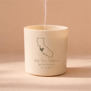 Puede incluir: Una vela encendida de color blanquecino con un gráfico del contorno del estado de California y un corazón. El texto "Big Sur, California" está debajo del gráfico, junto con los nombres "Bridget & Mark" y la fecha "07.13.2025".