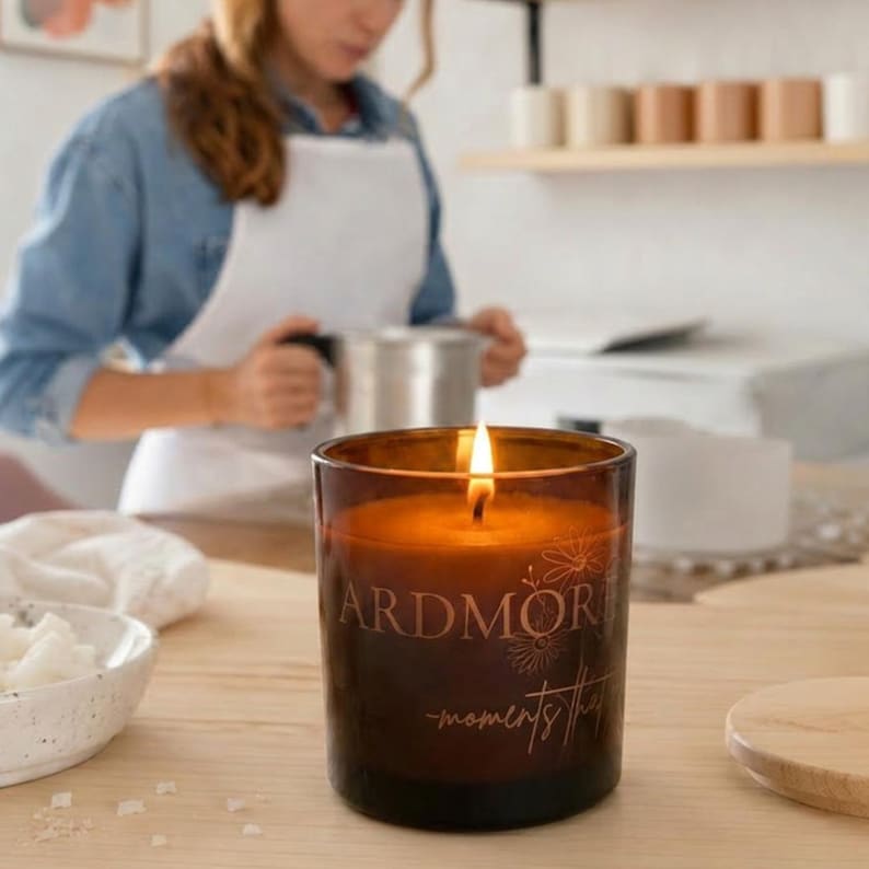 以下が含まれることがあります： 火のついた芯がある円筒形のアンバーガラス製キャンドルホルダー。ガラス容器の前面には、細い明るい色のフォントで「ARDMORE」および「moments that」という文字と、繊細な線画のフラワーイラストが描かれています。