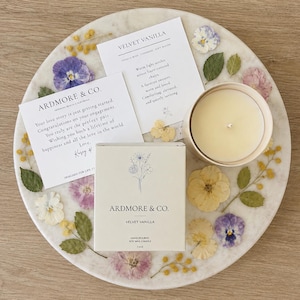 以下が含まれることがあります： セラミック容器に入った白いキャンドル、Ardmore & Co. Velvet Vanillaとラベルが貼られた長方形の箱、そして押し花で飾られた丸い大理石のトレイの上に置かれた2枚の印刷されたカード。箱には花のイラストとテキストの詳細が記載されています。