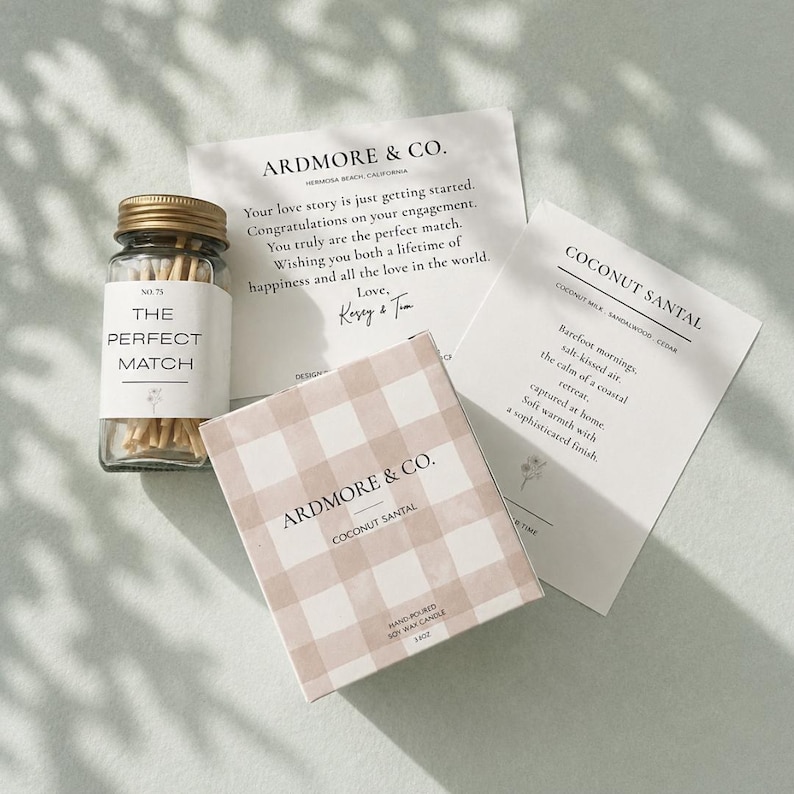 以下が含まれることがあります： The Perfect Matchと書かれたラベル付きのマッチのガラス瓶、Ardmore & Co. Coconut Santalと書かれたベージュと白のギンガムチェック柄の四角い箱、そして婚約と香りに関する黒いテキストが印刷された2枚の白いカード。