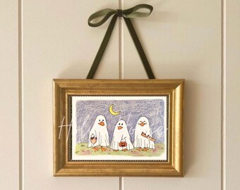 May include: A framed Halloween-themed artwork featuring three cartoon ghosts with orange beaks and feet, holding a basket, a pumpkin, and a treat. The artwork is set against a blue night sky with a crescent moon and is framed in a gold-colored frame, suspended by a green ribbon.