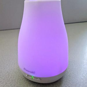 May include: A white and purple essential oil diffuser with the text "HomeweekS" on the front. The diffuser has a rounded shape and a green light indicating it is on. The diffuser is designed to release essential oils into the air.