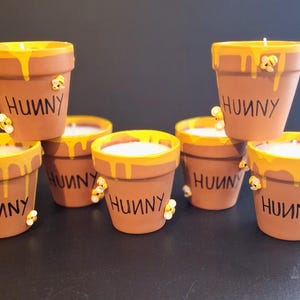 May include: A collection of terracotta-colored flower pot candles with yellow wax and honey-like drips. Each pot has the word "HUNNY" printed on it and is decorated with small bee embellishments. The candles are arranged on a dark surface.