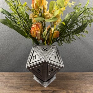 May include: A silver, 20-sided die shaped vase with a floral arrangement of yellow and orange flowers. The vase has numbers 3, 7, 11, 20, and 2 on the visible sides.