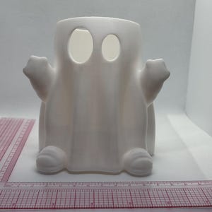 May include: White ceramic ghost figurine with two eye holes. The ghost has a rounded body and arms, and a pointed top.