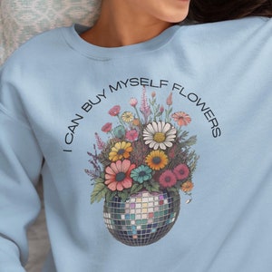 May include: A light blue sweatshirt with the text "I can buy myself flowers" in a circle around a bouquet of colorful flowers in a disco ball vase.