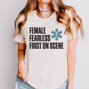 May include: A cream-coloured t-shirt with the words "FEMALE FEARLESS FIRST ON SCENE" in black block letters. A blue medical symbol is to the right of the text. The shirt is a crew neck and has short sleeves.