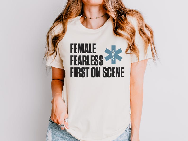 May include: A cream-coloured t-shirt with the words "FEMALE FEARLESS FIRST ON SCENE" in black block letters. A blue medical symbol is to the right of the text. The shirt is a crew neck and has short sleeves.