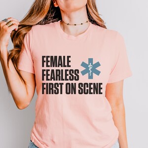 May include: A pale pink t-shirt with the text "FEMALE FEARLESS FIRST ON SCENE" in black. A blue medical symbol is next to the text. The t-shirt has a crew neck.