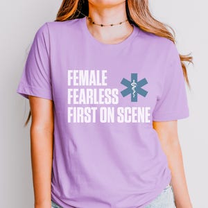 May include: A lilac t-shirt with the text "FEMALE FEARLESS FIRST ON SCENE" in white. A blue medical symbol is to the right of the text. The shirt has a crew neck design.