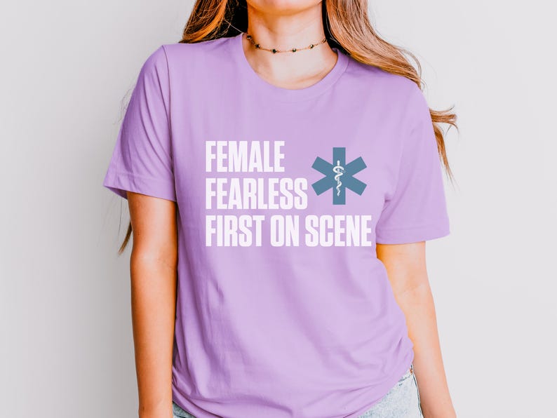 May include: A lilac t-shirt with the text "FEMALE FEARLESS FIRST ON SCENE" in white. A blue medical symbol is to the right of the text. The shirt has a crew neck design.