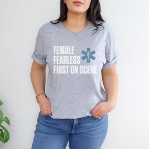 May include: A light grey t-shirt with rolled sleeves. The shirt displays the text "FEMALE FEARLESS FIRST ON SCENE" in white, and a teal medical symbol. The shirt is worn with blue jeans.