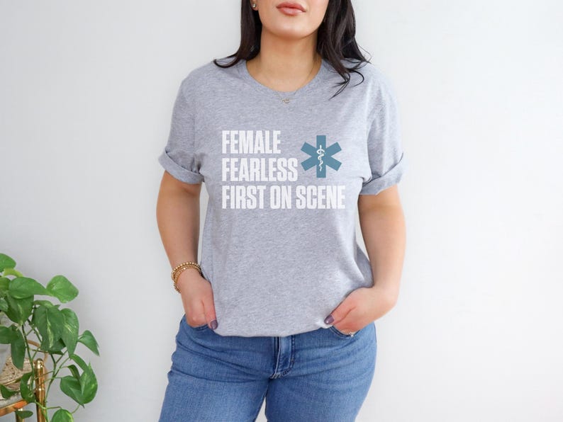 May include: A light grey t-shirt with rolled sleeves. The shirt displays the text "FEMALE FEARLESS FIRST ON SCENE" in white, and a teal medical symbol. The shirt is worn with blue jeans.