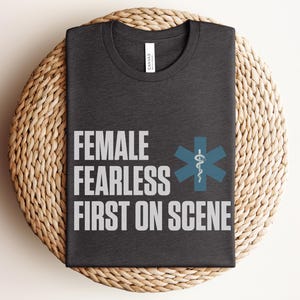 May include: Dark grey t-shirt with the words "FEMALE FEARLESS FIRST ON SCENE" in white block letters. A blue medical symbol is to the right of the text. The shirt is folded on a woven, circular surface.