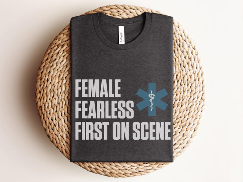 May include: Dark grey t-shirt with the words "FEMALE FEARLESS FIRST ON SCENE" in white block letters. A blue medical symbol is to the right of the text. The shirt is folded on a woven, circular surface.