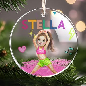 May include: A round, clear acrylic ornament featuring the name STELLA in colorful block letters. The design includes a cartoon illustration of a child in a pink sports bra and lime green shorts, surrounded by a pink heart, music note, and lightning bolt.
