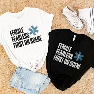 Two t-shirts, one white and one black, with the text 'Female Fearless First On Scene' and a blue medical star symbol.