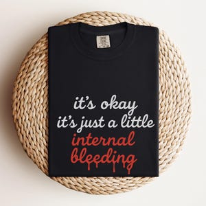 May include: Black t-shirt with the text "it's okay it's just a little internal bleeding" in white and red lettering. The shirt is displayed on a round, woven, beige placemat. The shirt has a tag on the upper right side.