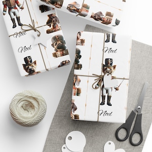 Puede incluir: Papel de regalo blanco con un patrón repetido de perros vestidos de cascanueces. El papel tiene un borde dorado y la palabra "Noël" está impresa en el papel.