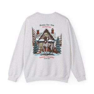 Op de afbeelding: Een grijs sweatshirt met een grafische print van een hond die voor een peperkoekhuis staat. De tekst "Home for the Holidays" en "Santa's Favorite Good Boy" staat op het sweatshirt gedrukt.