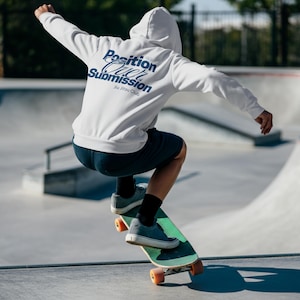Può includere: Felpa con cappuccio bianca con la scritta "Position Over Submission Jiu Jitsu Club" in blu navy. La persona sta andando sullo skateboard con una tavola verde e ruote arancioni. La persona indossa pantaloncini blu navy e scarpe da ginnastica grigie.