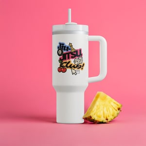May include: A white tumbler with a handle and a straw, featuring a colorful "Jiu Jitsu Club!" graphic with cherries, dice, and a chess piece. A slice of pineapple rests beside the tumbler against a pink background.