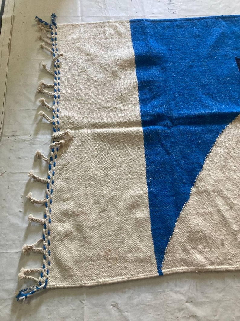 Peut inclure: Un tapis tiss&eacute; blanc et bleu avec des franges sur un c&ocirc;t&eacute;. Le tapis a un motif g&eacute;om&eacute;trique avec un grand triangle bleu.