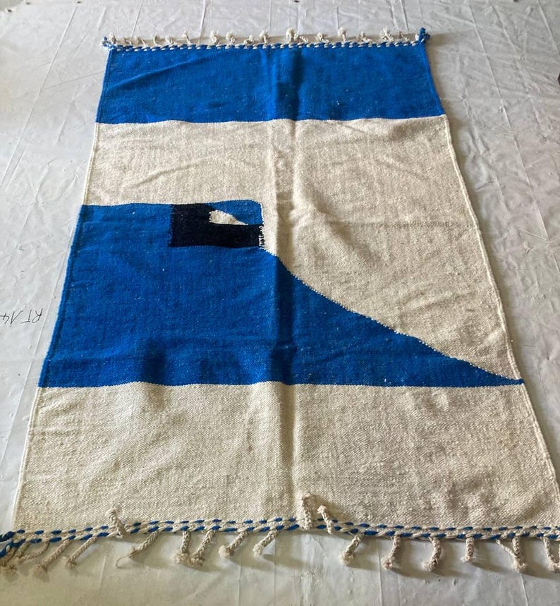Peut inclure: Un tapis tiss&eacute; blanc et bleu avec un motif g&eacute;om&eacute;trique. Le tapis a des franges sur tous les c&ocirc;t&eacute;s.