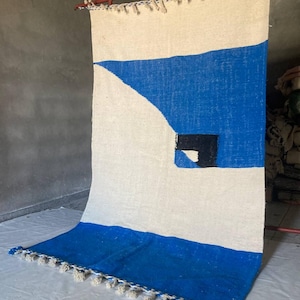 Peut inclure: Un tapis tiss&eacute; blanc et bleu avec un motif g&eacute;om&eacute;trique. Le tapis a une bordure &agrave; franges et un carr&eacute; noir au centre.