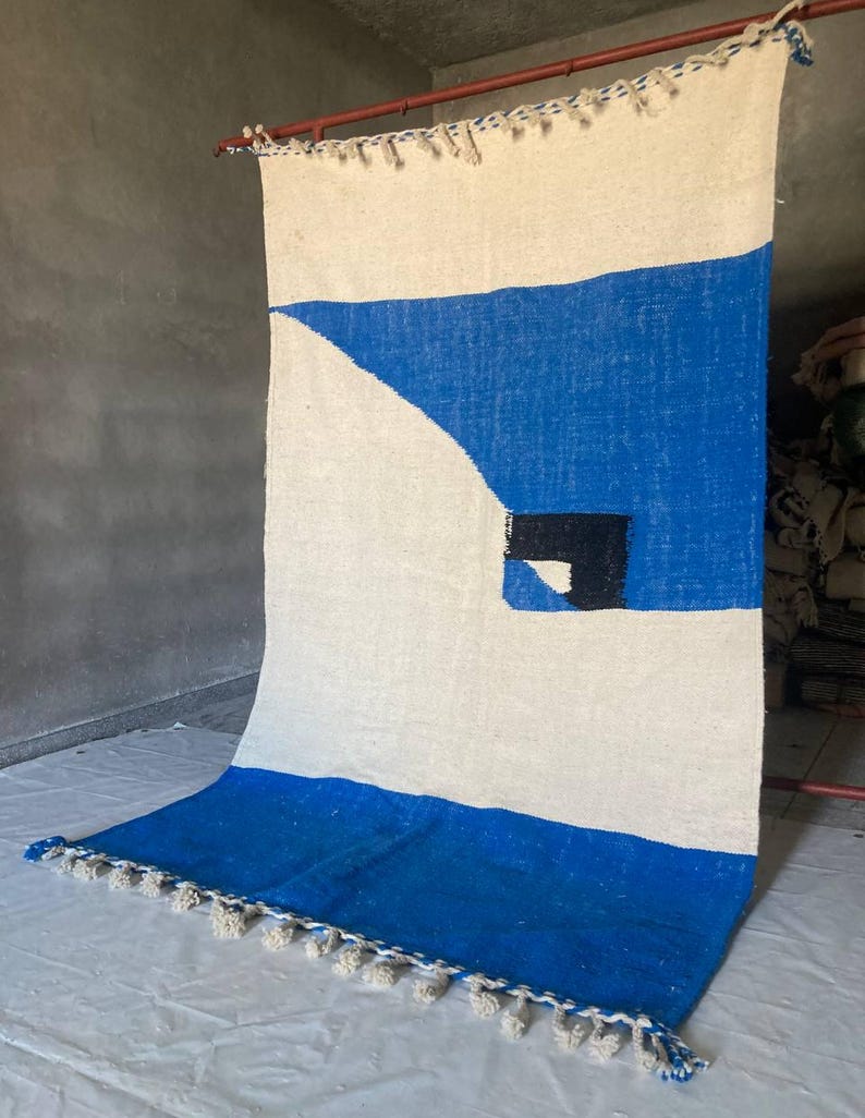 Peut inclure: Un tapis tiss&eacute; blanc et bleu avec un motif g&eacute;om&eacute;trique. Le tapis a une bordure &agrave; franges et un carr&eacute; noir au centre.
