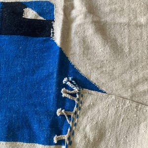Peut inclure: Une couverture en laine tiss&eacute;e bleue et blanche avec des franges sur les bords. La couverture a un motif g&eacute;om&eacute;trique et est faite d'un mat&eacute;riau naturel doux.