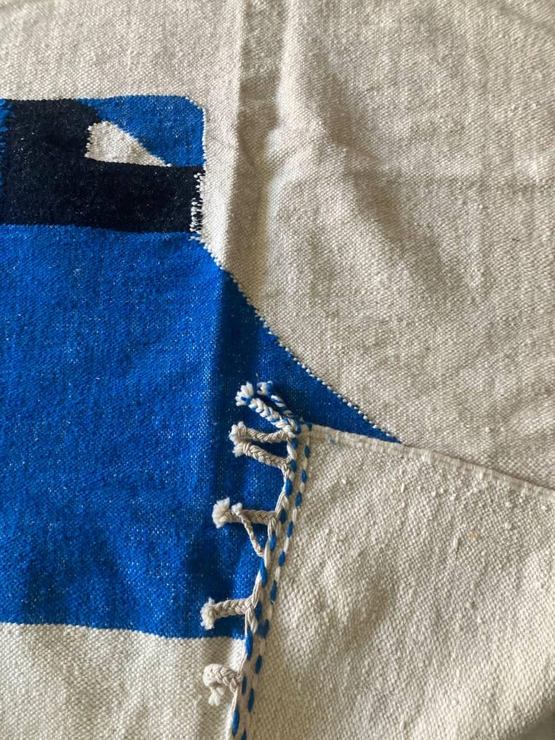 Peut inclure: Une couverture en laine tiss&eacute;e bleue et blanche avec des franges sur les bords. La couverture a un motif g&eacute;om&eacute;trique et est faite d'un mat&eacute;riau naturel doux.