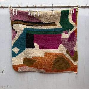 May include: A colorful, abstract woven wall hanging. The textile features a geometric design with a mix of colors, including teal, fuchsia, orange, green, and cream. The top edge has small white tassels. The wall hanging is suspended from a metal rod.