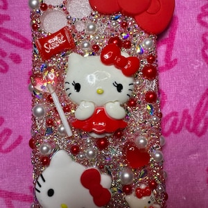 May include: A pink and red Hello Kitty phone case with rhinestones, pearls, and a red bow. The case has a Hello Kitty character with a red dress and a lollipop. The case also has a red candy bar with the word "Sweet" on it.