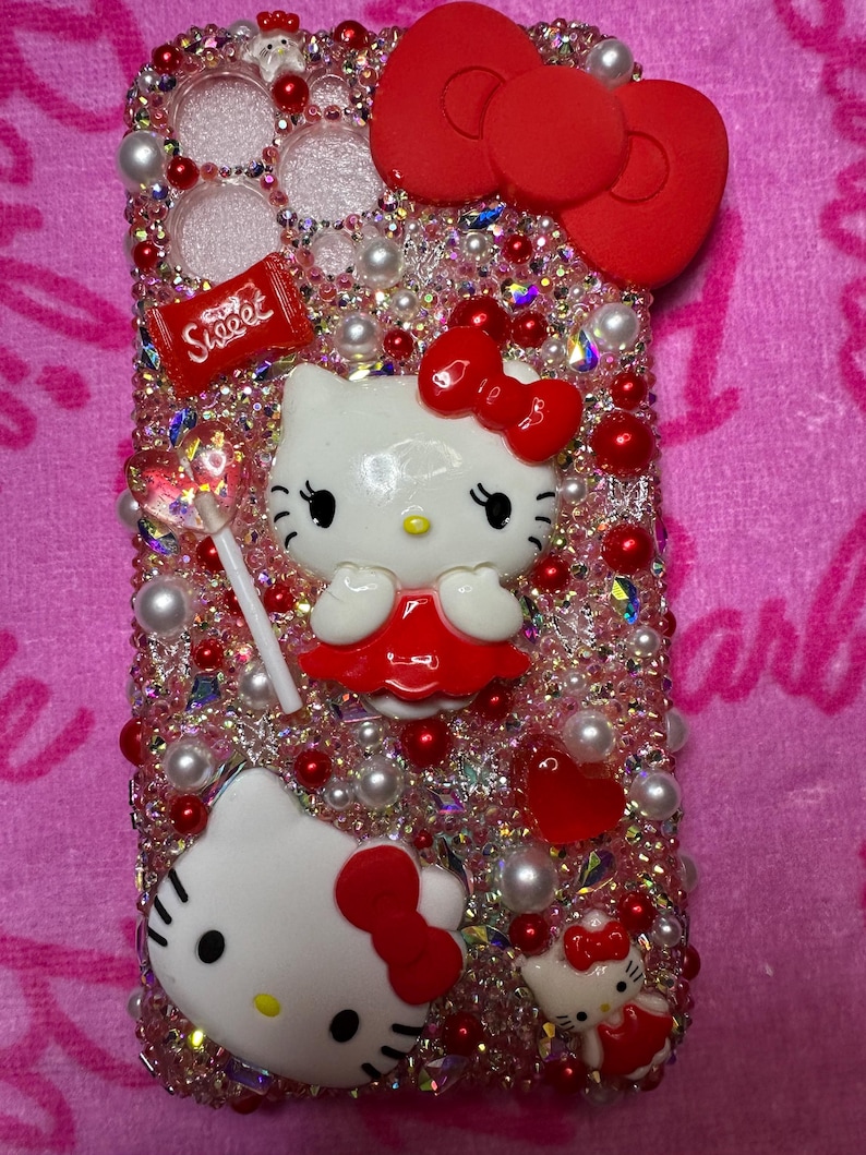 May include: A pink and red Hello Kitty phone case with rhinestones, pearls, and a red bow. The case has a Hello Kitty character with a red dress and a lollipop. The case also has a red candy bar with the word "Sweet" on it.