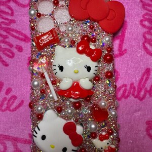 May include: A pink and red Hello Kitty phone case with rhinestones, pearls, and a red bow. The case features a Hello Kitty character holding a lollipop with the word "Sweet" on it.