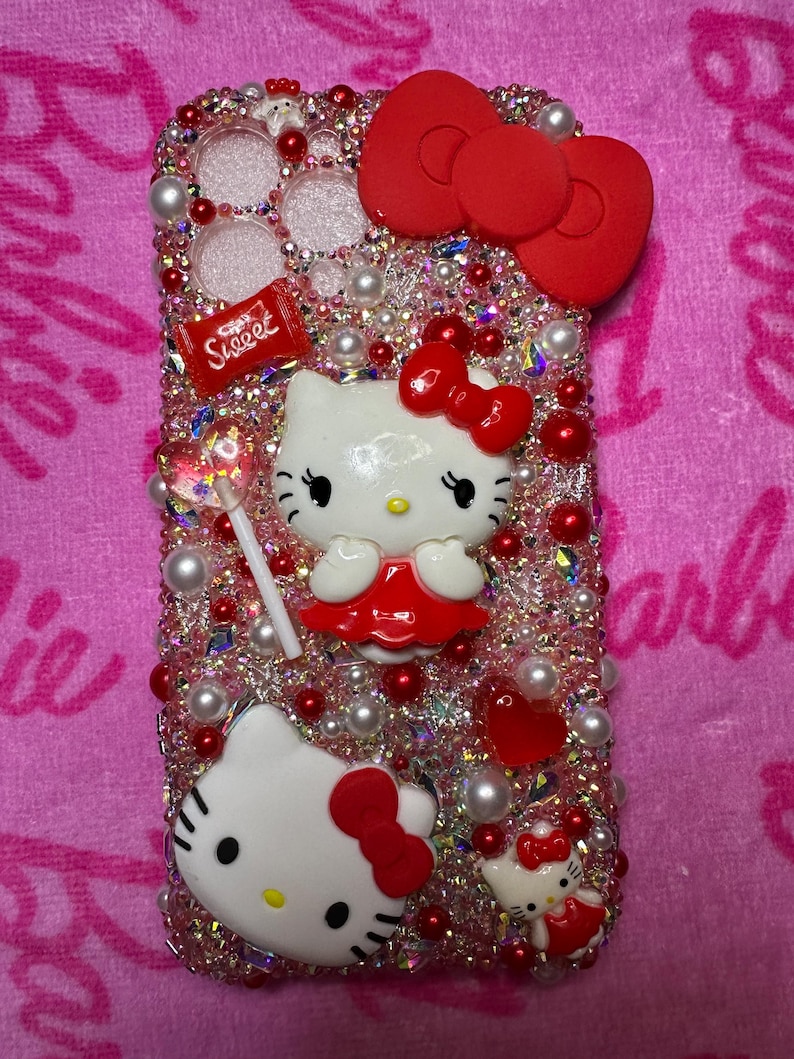 May include: A pink and red Hello Kitty phone case with rhinestones, pearls, and a red bow. The case features a Hello Kitty character holding a lollipop with the word "Sweet" on it.