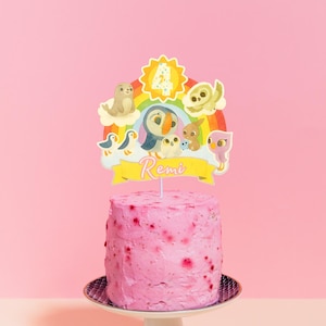 Könnte beinhalten: Ein rosa glasierter Kuchen mit einem dekorativen Aufsatz mit einem Regenbogen, Zeichentricktieren und der Zahl 4. Der Kuchen steht auf einem weißen Tortenständer vor einem rosa Hintergrund. Der Name "Remi" steht auf dem Aufsatz.