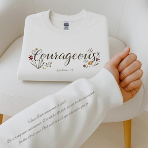 May include: A white sweatshirt with the word "Courageous" in a script font, adorned with floral accents. Below the word is "Joshua 19." The sleeve displays a quote: "Have I not commanded you? Be strong and courageous..." A comfortable, inspirational garment.