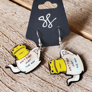 May include: A pair of silver dangle earrings featuring a yellow frog sitting in a white teapot with the text "Ain't that just the way" on the side.
