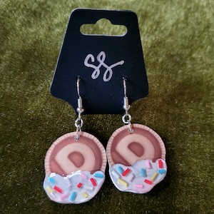May include: A pair of dangle earrings featuring a donut-shaped charm with white frosting and colorful sprinkles. The donut charm is brown and has a white 'D' in the center.