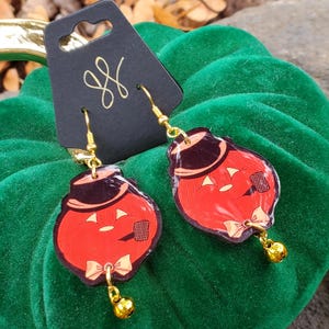 May include: A pair of dangle earrings featuring a red pumpkin character wearing a black top hat and a tan bow tie, with a small gold bell attached to the bottom. The earrings are displayed on a black card with a gold logo at the top.