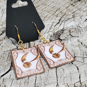 May include: Gold earrings with a white background featuring a golden snitch and a decorative border. The earrings are shaped like a rectangle.