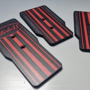 Puede incluir: Tres objetos rectangulares negros y rojos con esquinas redondeadas. Los objetos tienen rayas verticales rojas. Un objeto tiene la palabra "Peterbilt" en rojo. Cada objeto tiene dos agujeros.