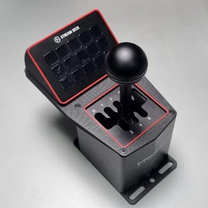 Puede incluir: Stream Deck negro y rojo con una palanca de cambios negra. El Stream Deck tiene 15 botones negros y el texto "STREAM DECK". La palanca de cambios tiene una bola negra en la parte superior y un patrón de engranajes con números.