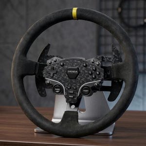 Può includere: Un volante da corsa nero con una texture simile alla pelle scamosciata, dotato di un segno centrale giallo sulla parte superiore. Il mozzo centrale mostra il testo MOZA RACING, vari pulsanti circolari, un pad direzionale e leve del cambio geometriche.